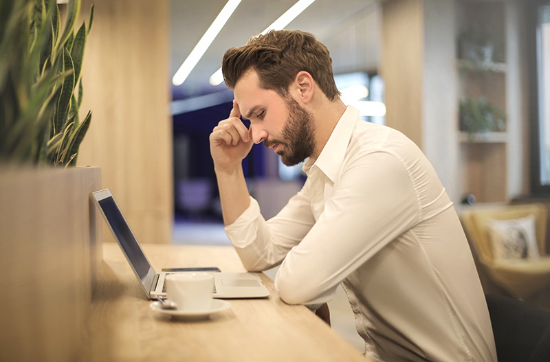 A man drinking coffee sits and studies his laptop with a thoughtful expression.