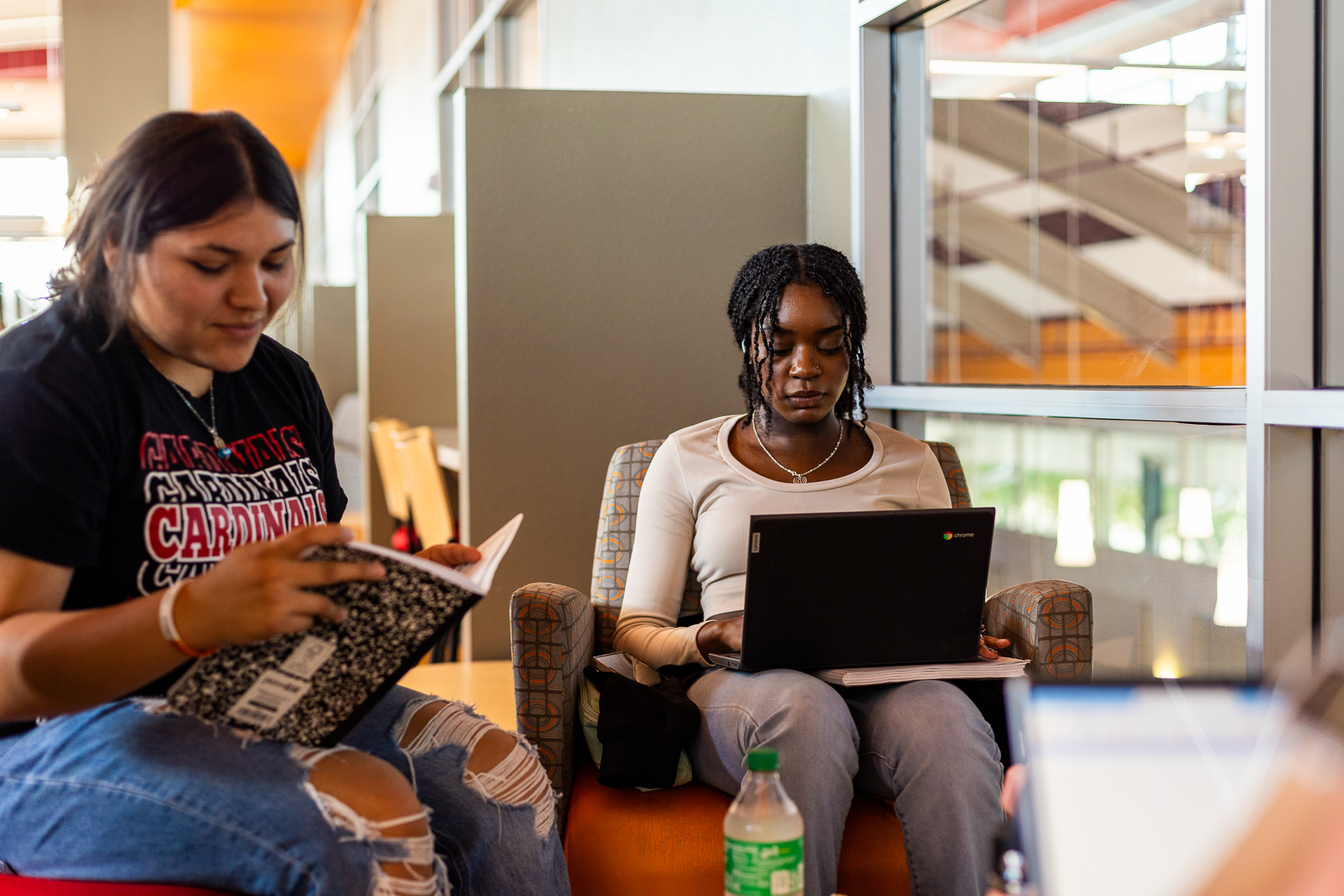 Two young women sitting together, one reading a book and the other using a laptop