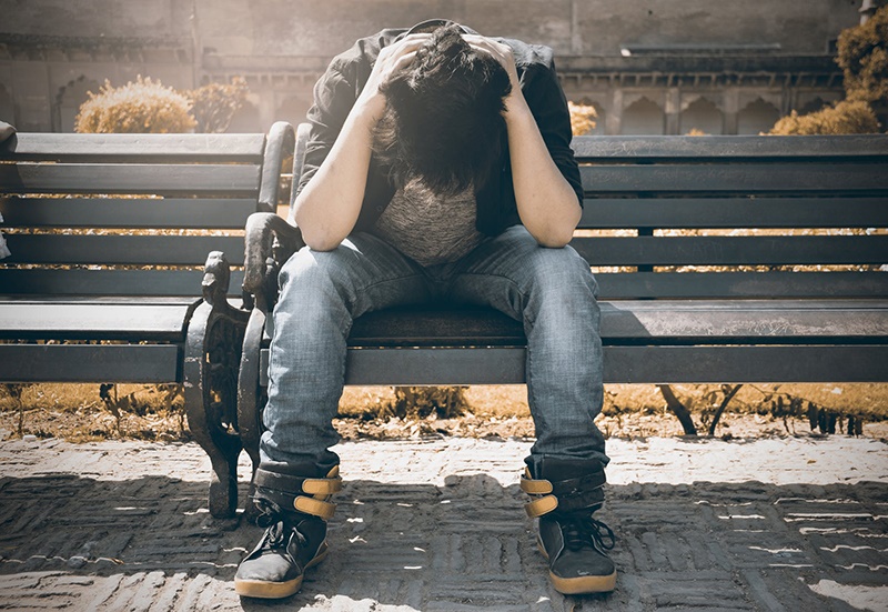 A man sits alone on a bench, hunched forward with hands on their head.