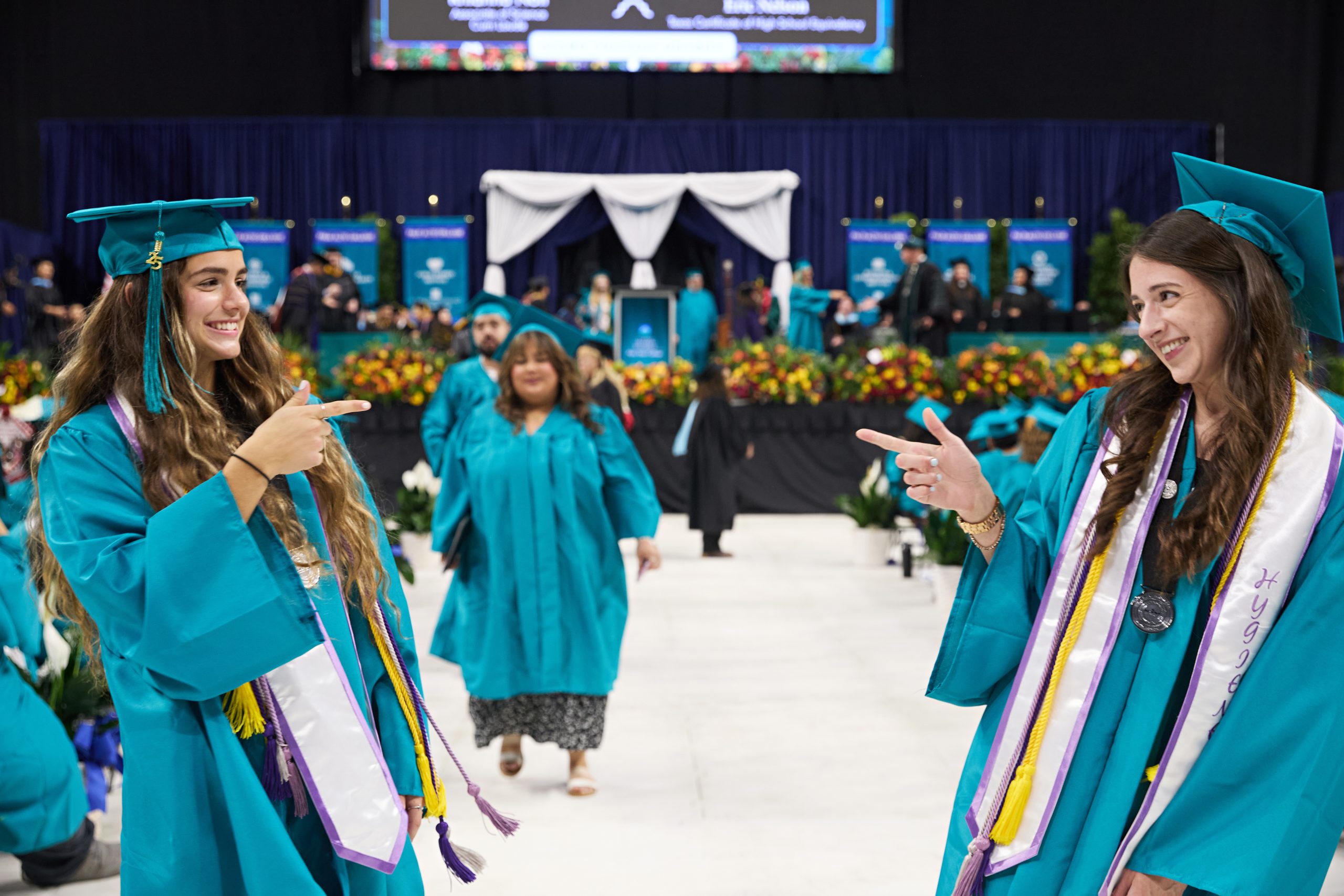 Two young women in graduation regalia smile and point at one another.