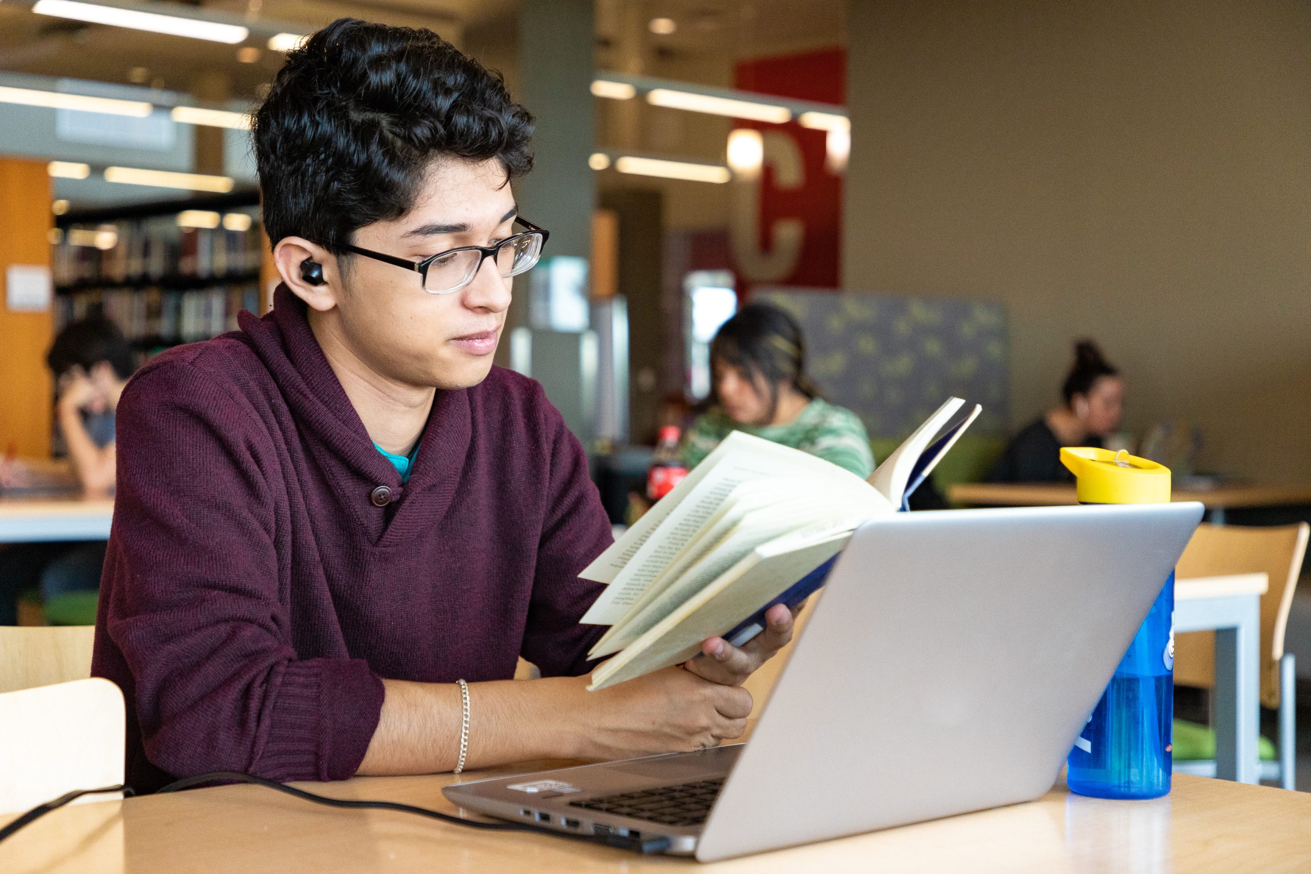 A man reads a book in front of his laptop, with library bookshelves in the background.