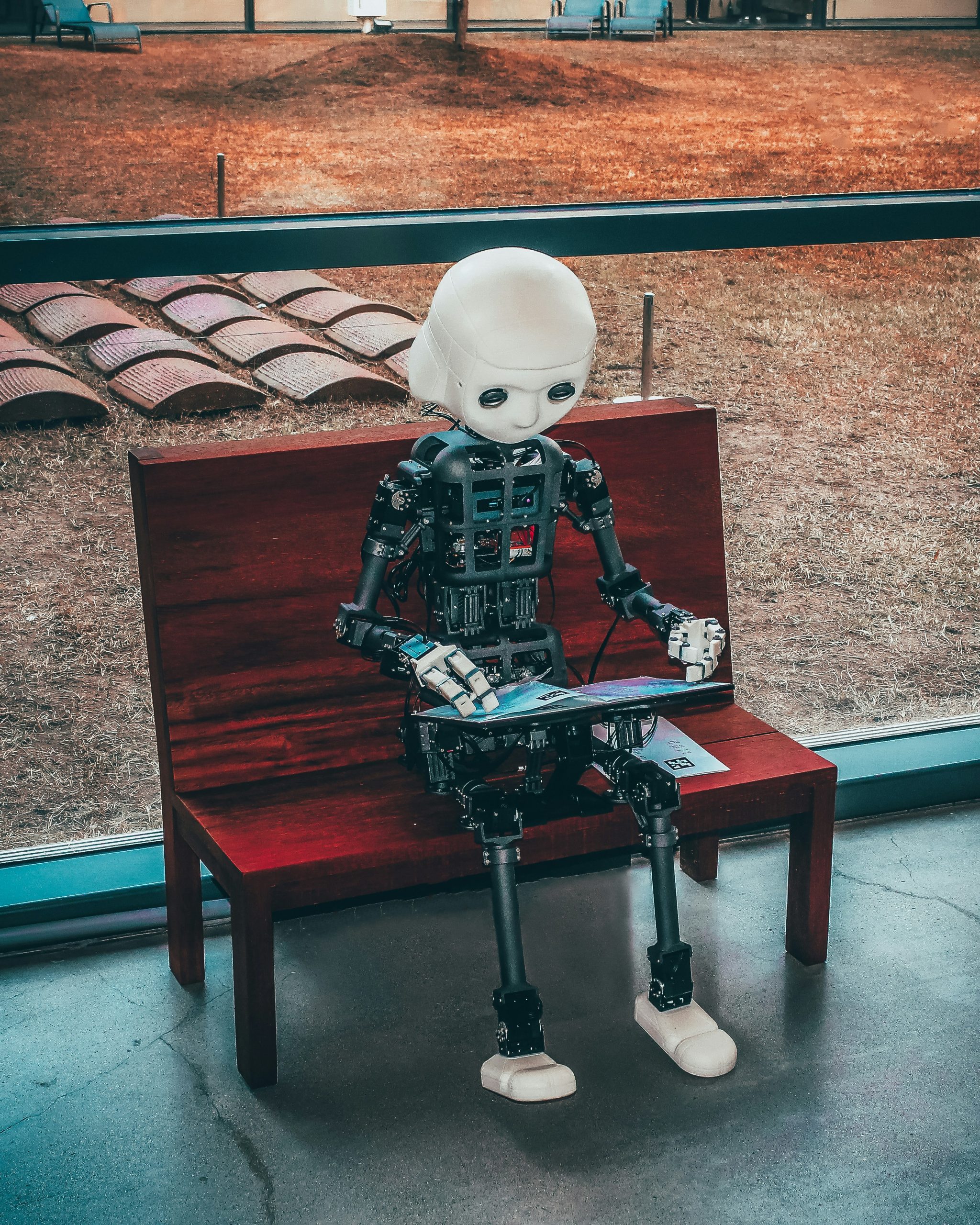 A robot configured to look like a human sits on a bench and reads.